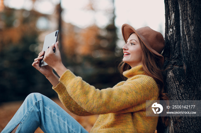 Young woman model sitting with tablet pc in autumn park with yellow foliage maple leaves. Fall season fashion