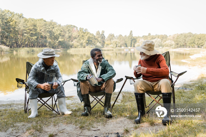 Cheerful multicultural men in fishing outfit holding thermo cups near lake