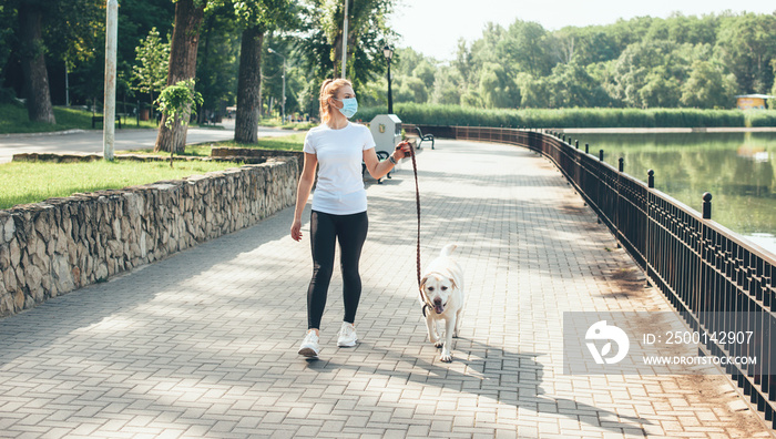 Full sized photo of a caucasian woman with blonde hair and medical mask on face walking with her golden dog