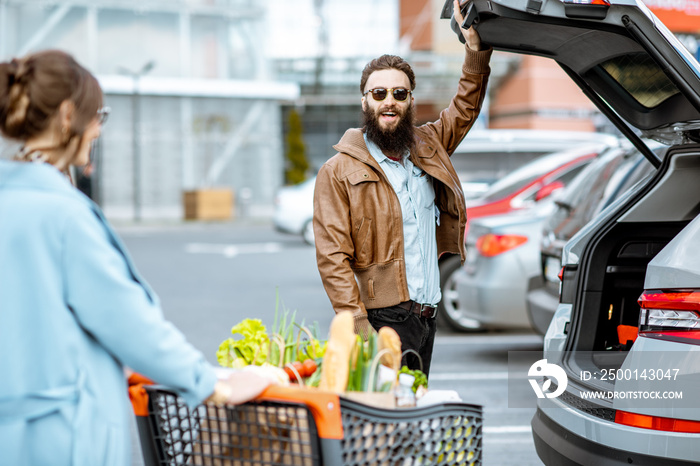 Woman riding shopping cart full of food with man waiting near the car on the outdoor parking