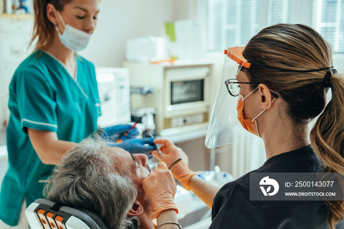 Unrecognizable male patient sitting on a dentist’s chair and getting dental restoration, while female dental assistant giving dental instrument to dentist in a dental clinic