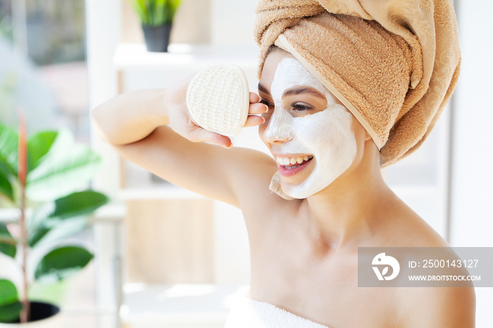 Young happy woman in towel, applying facial clay Mask in stylish bathroom.