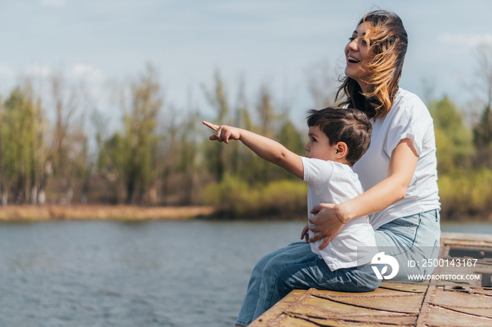 Cute kid pointing with finger while sitting near happy mother and lake