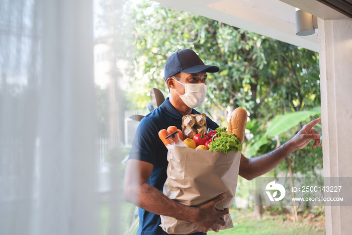 African American delivery man wearing protective face mask holding grocery paper bag of Healthy Food with fresh vegetables, milk, bread.