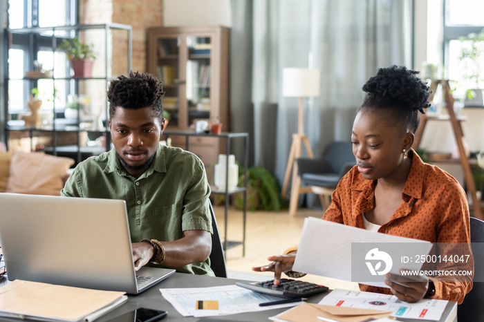 Concentrated young African American couple sitting at table in living room and calculating tax bills