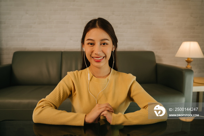 Portrait of smiling young successful Asian woman on video call using headphones sitting focused and concentrated while working late from home at night