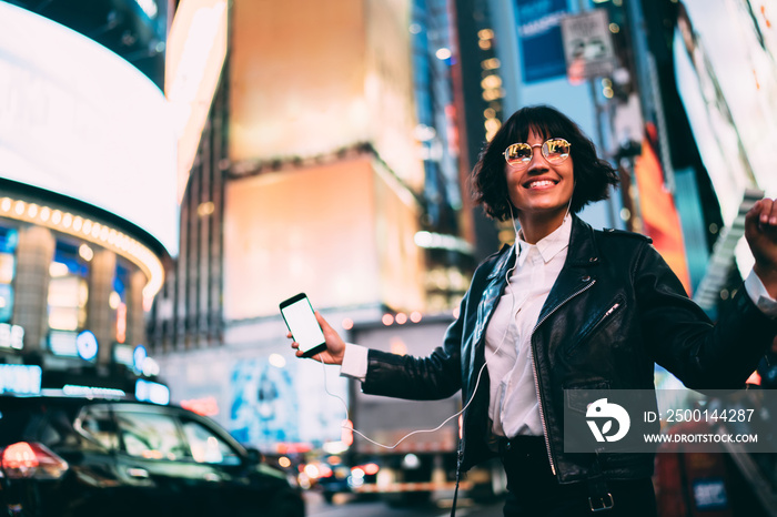 Cheerful woman in fashionable clothing listening positive music for relaxing in evening city, dancing teenager in electronic headphones holding blank smartphone technology and recreating outdoors