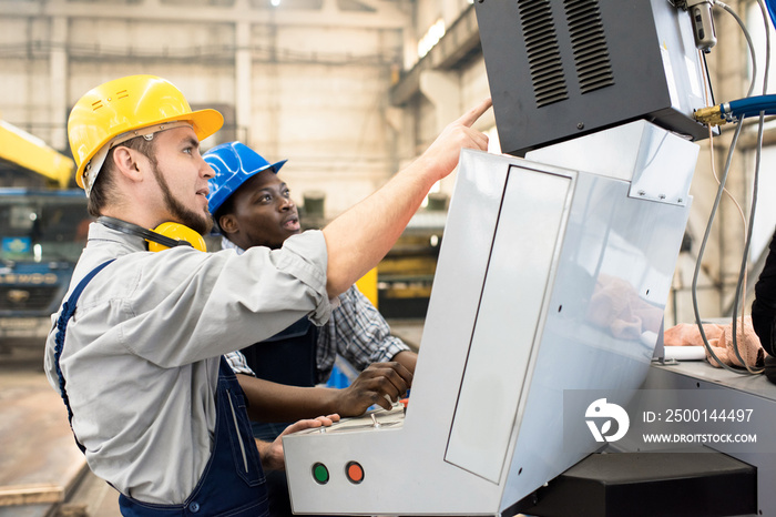 Multi-ethnic team of machine operators wearing hardhats and overalls discussing working process while standing in front of CNC, interior of production department on background