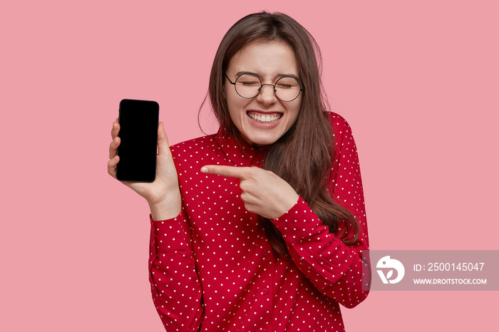 Optimistic Caucasian woman points at new smart phone for advertising, shows blank screen, likes multifunctional gadget, wears red shirt, poses over pink background. People and promotion concept