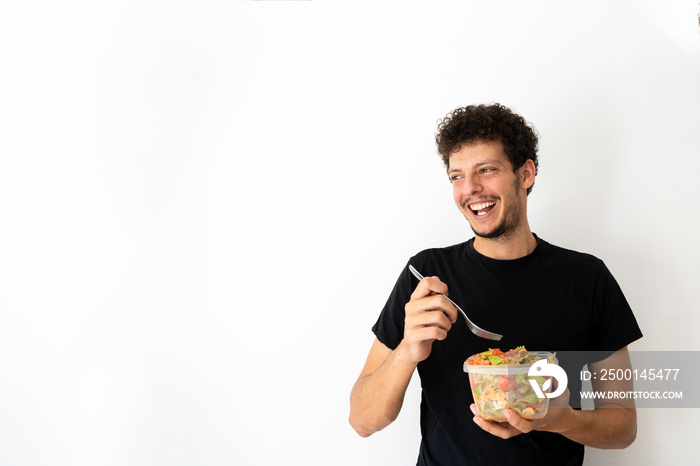 Happy young caucasian man eating and holding a healthy mediterranean pasta salad tupperware