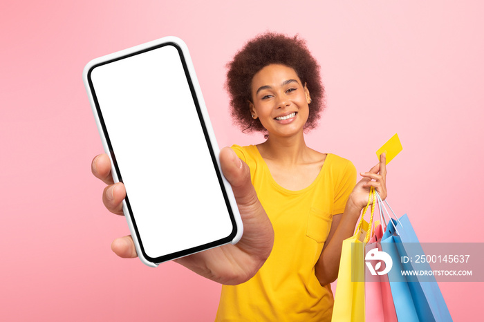 Cheerful young african american curly female with bags with purchases, credit card show smartphone with blank screen