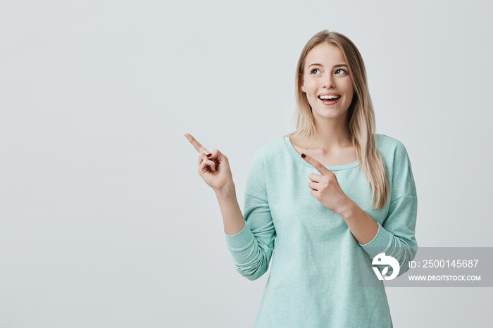 Happy surprised blonde young female smiling broadly at camera, pointing fingers away, showing something interesting and exciting on studio wall with copy space for your text or advertising content