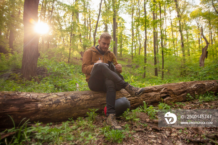 one young man, in forest, holding a flower in his hands, writing down his notes on it.