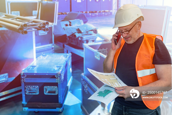 Human in a building uniform. Engineer next to musical equipment. Concept - installation of sound equipment. Concept - electrician talking on phone. Drawings of concert venue in the hands of a person