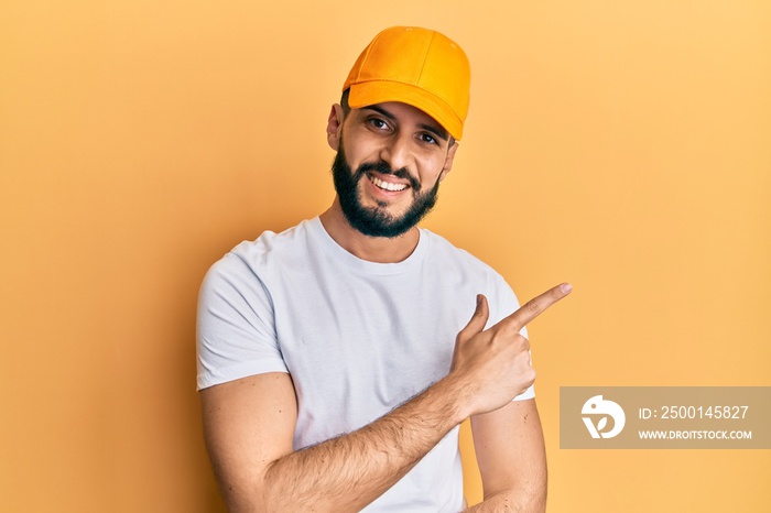 Young man with beard wearing yellow cap smiling cheerful pointing with hand and finger up to the side