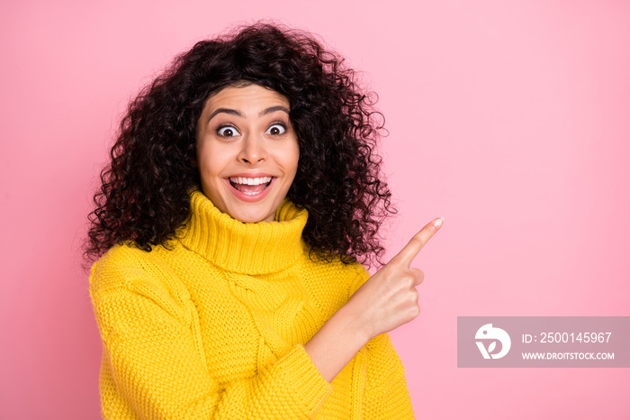 Photo portrait of surprised curly girl in knitted sweater pointing finger blank space isolated on pastel pink background