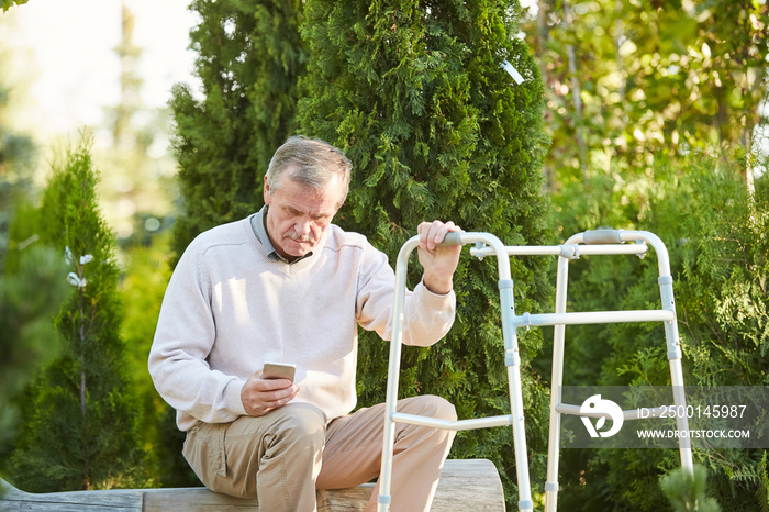 Portrait of senior man with walker resting on park bench using smartphone, copy space