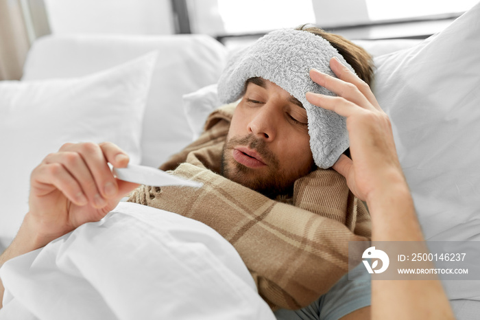 people, health and fever concept - sick man with cold compress on his forehead measuring temperature by thermometer lying in bed at home