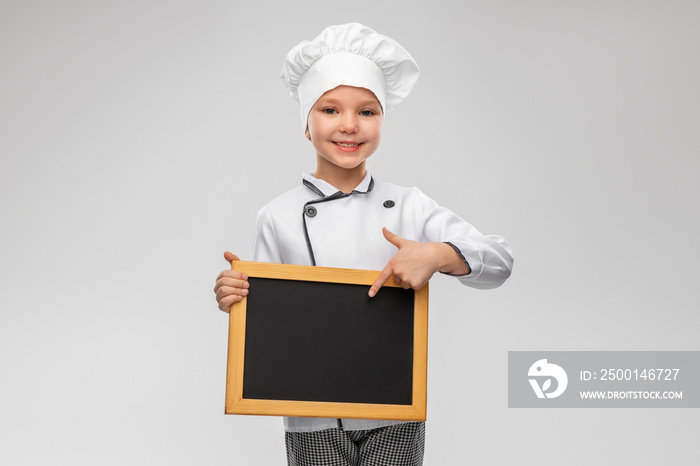 cooking, culinary and profession concept - happy smiling little girl in chef’s toque and jacket holding chalkboard over grey background