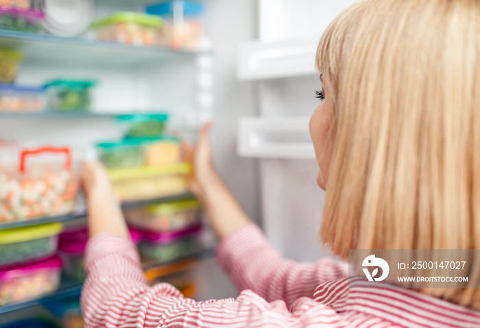 Young woman puts containers with frozen mixed vegetables into freezer.