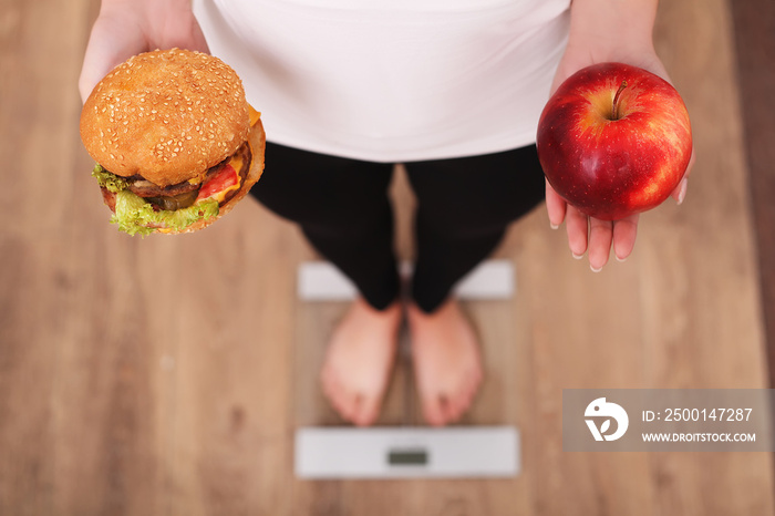 Diet. Woman Measuring Body Weight On Weighing Scale Holding Burger and apple. Sweets Are Unhealthy Junk Food. Dieting, Healthy Eating, Lifestyle. Weight Loss. Obesity. Top View