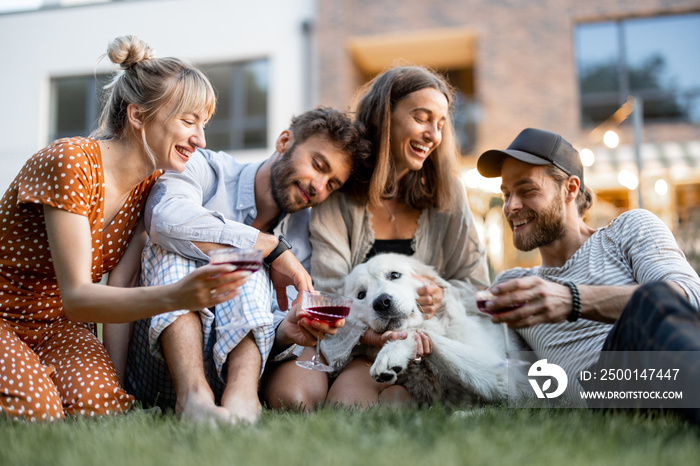 Young friends having fun playing with a dog and drinking wine, sitting on the green lawn at backyard of the country house in the evening