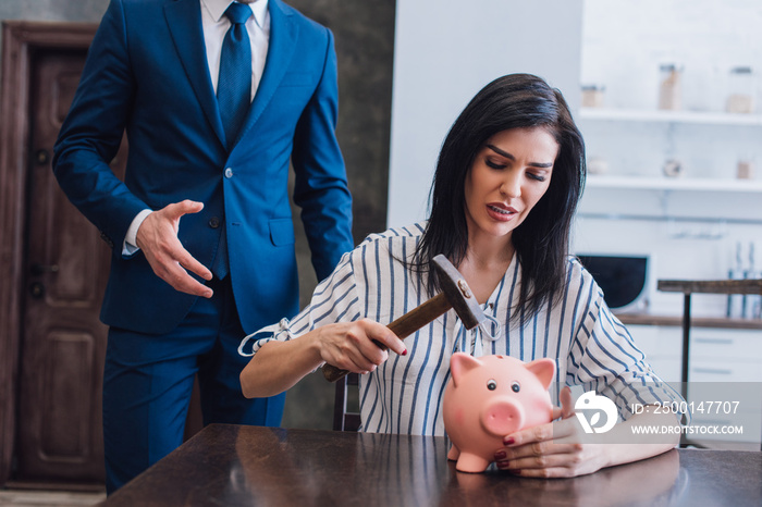 Worried woman holding hammer above piggy bank at table near collector with outstretched hand in room