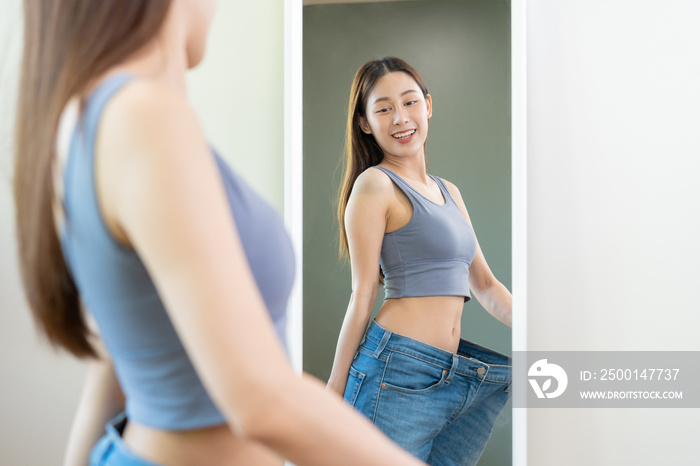 Shape slender, thin waist, attractive slim asian young woman, beautiful girl hand show shape her weight loss, wearing in big, large or over size jeans and looking into mirror. People body fit healthy.