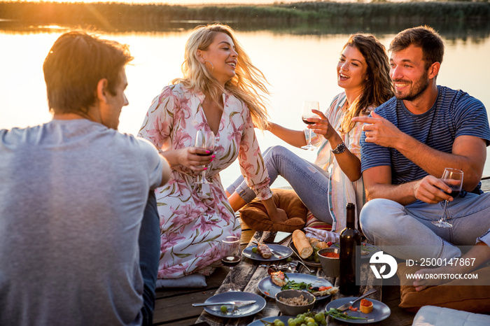 Group of friends having fun on picnic near a lake, sitting on pier eating and drinking wine.