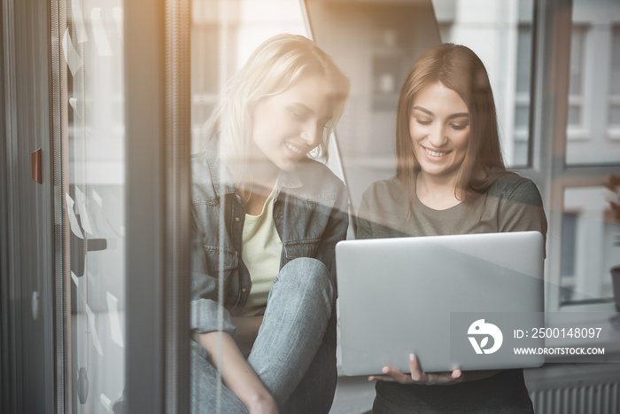 Young girl sitting on windowsill in office. Colleague standing next to her with laptop in hand. Both are looking at notebook screen with satisfaction