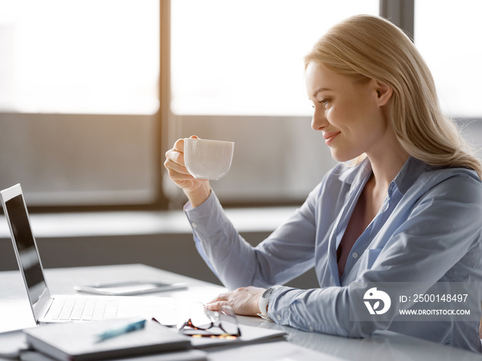 Happy young woman is drinking hot beverage at workplace. She is looking at cup with joy and smiling