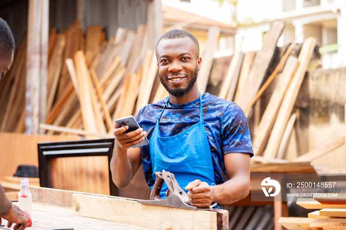 image of cheerful african man with a working tool and a smart phone- young black entrepreneur in a workshop