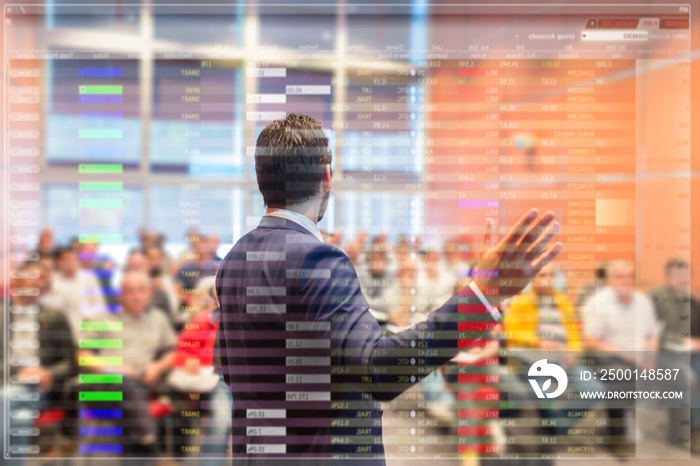 Speaker at business conference with public presentations. Audience at the conference hall. Trough the transparent presentation screen view. Charts and tables can be seen in forground.