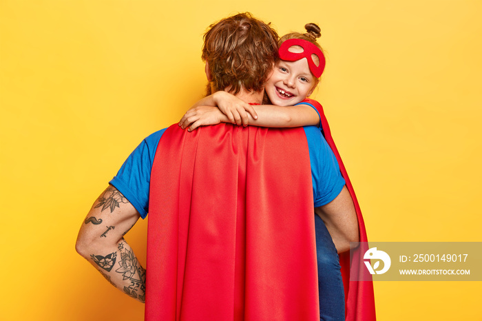 My lovely super dad. Affectionate small child cuddles father with love, wears red mask, dressed in superhero costumes, confident in defense and dads strength, play together, isolated on yellow wall