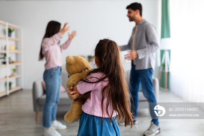 Family quarrels concept. Little girl suffering from family quarrels, cuddling her teddy bear and turning back to parents