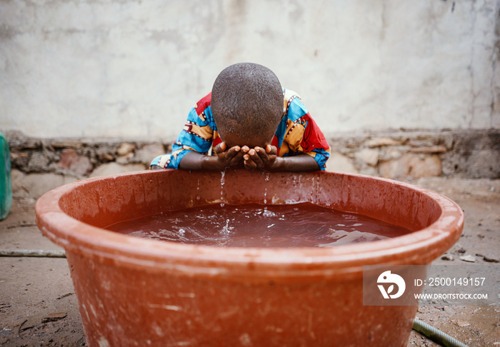 Little black african boy washing his face over a huge brown plastic basin with clean fresh water