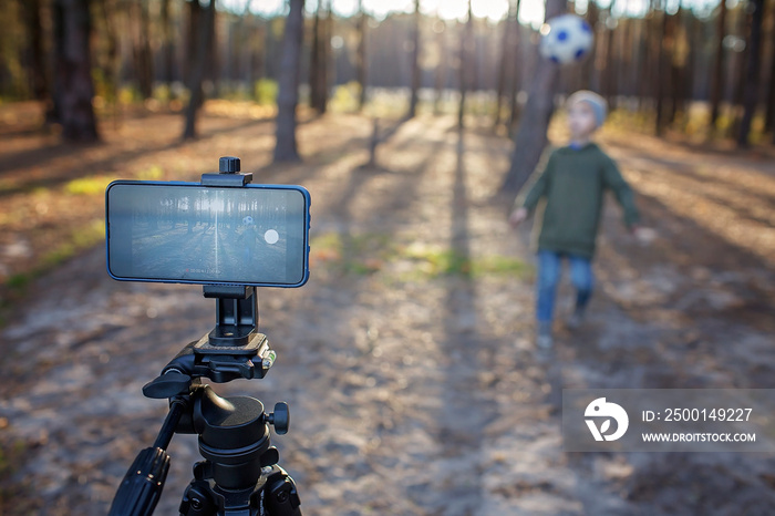 Little boy, young football player, hitting the ball and recording viral video film with smartphone to share in social media in autumn forest, sport blogger, outdoor lifestyle, healthy active life