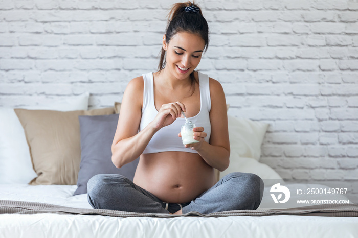 Beautiful young pregnant woman eating yogurt while sitting on the bed at home.