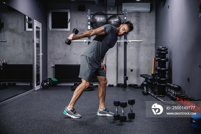 Hand training with sports equipment. Full body portrait of a handsome and muscular man in the gym doing triceps exercises with dumbbells. Modern gym with a dark atmosphere, sports life