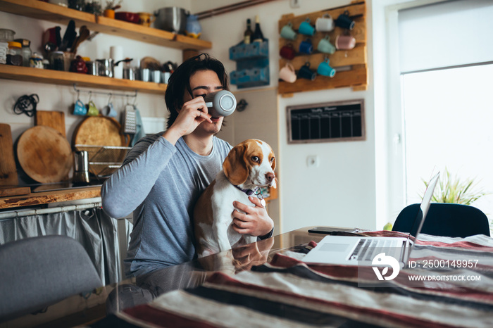 young man holding his dog in nap while using laptop in kitchen