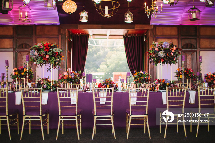 Purple banquet table in tropical style, wedding decor. Purple tablecloth, exotic flowers. There are geometric and classical chandeliers on the ceiling and panoramic windows in the background.