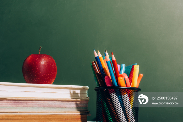 Pen holder with color pencils and felt pens and ripe apple on books near green chalkboard