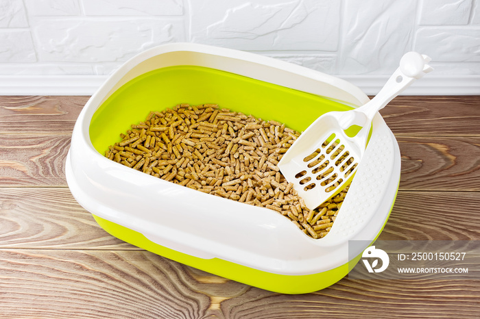 High sided cat litter tray with wooden pellets and scoop on a brown wooden floor. New green cat box near the wall. Toilet for domestic pets