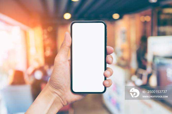 Man’s hand shows mobile smartphone with white screen in vertical position, Blurred or Defocus image of Coffee Shop or Cafeteria for use as Background vintage tone. - mockup template and clipping path