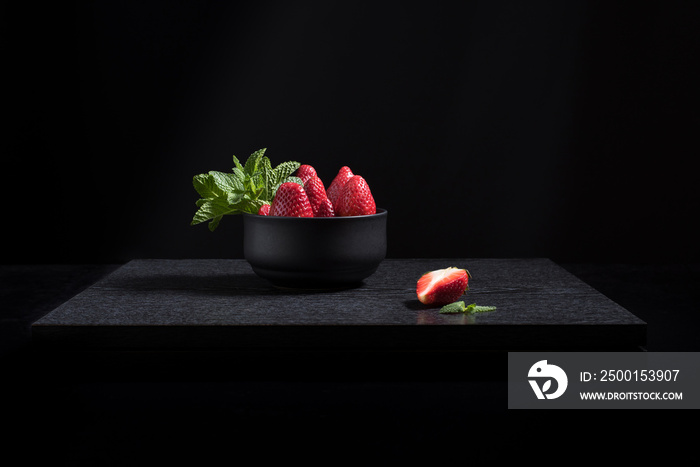 Fresas con hojas de menta en un recipiente negro sobre una mesa de madera negra y fondo oscuro