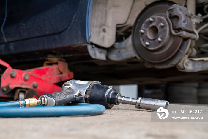 Pneumatic wrench tool on the asphalt and a car jack for lift up the body and changing the tire. Car without wheel.