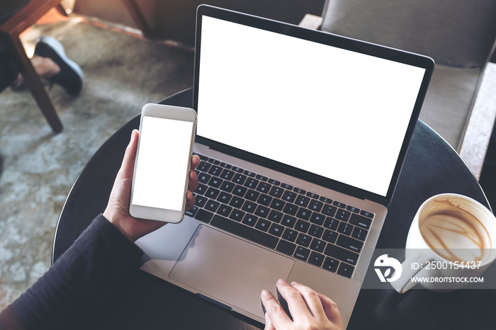 Top view mockup image of hands holding blank mobile phone while using laptop with blank white desktop screen on table