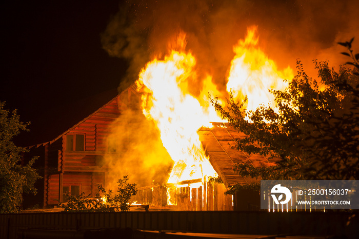 Burning wooden house at night. Bright orange flames and dense smoke from under the tiled roof on dark sky, trees silhouettes and residential neighbor cottage background. Disaster and danger concept.