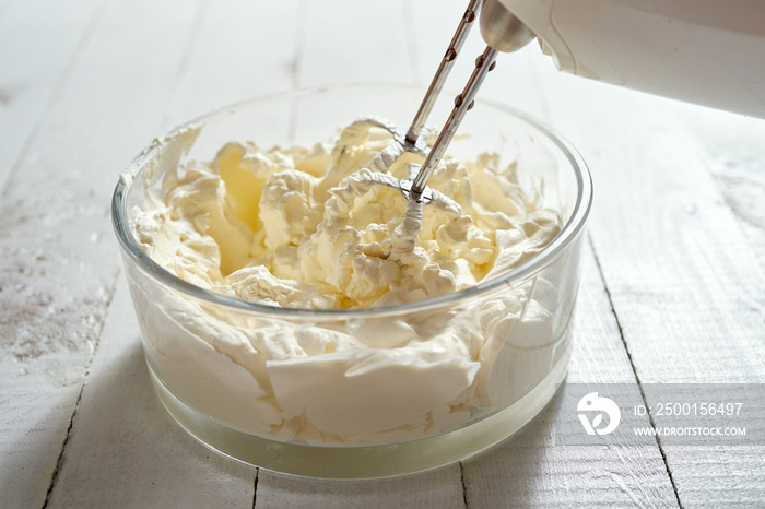 Whipped cream in a bowl and electric hand mixer on bright table.