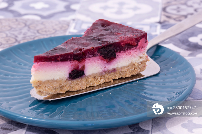 Slice of summer fruit cheesecake with a biscuit base served with a cake cutter utensil on a blue plate. Blue patterned tiled background